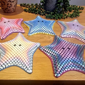 May include: Five decorative starfish in various colors, including pink, blue, and iridescent hues. Each starfish has a textured, scale-like pattern and two small, dark holes. They are arranged on a wooden surface.
