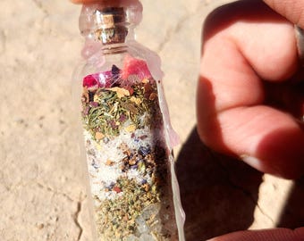 May include: A small, clear glass bottle filled with layers of dried herbs, crystals, and flower petals. The bottle is sealed with a cork and a clear wax. The contents are a mix of green, white, and pink. The bottle is held by a person's fingers.