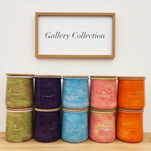 May include: A selection of brightly coloured ceramic jars with wooden lids. The jars are in shades of green, purple, blue, pink, and orange. Each jar has a unique design and text. A framed sign above reads "Gallery Collection."
