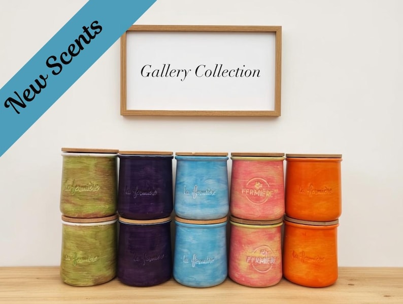 May include: Ten cylindrical ceramic jars with wooden lids arranged in two rows. The jars feature a textured, hand-painted finish in green, purple, blue, pink, and orange. Each jar displays the embossed text "La Fermi&egrave;re" and a framed sign reads "Gallery Collection" with a blue banner stating "New Scents".