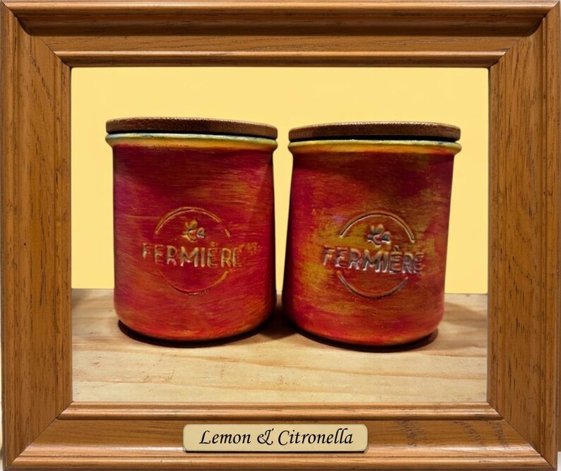 May include: Two cylindrical containers with a textured red and yellow finish, each featuring a wooden lid and the embossed text FERMIERE with a small bee icon. The containers are framed by a wooden border with a label reading Lemon & Citronella.