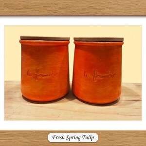 May include: Two cylindrical orange ceramic canisters with flat wooden lids. Each canister features the embossed cursive text "la fermi&egrave;re" on the front surface. The containers have a smooth, matte finish and a slightly tapered shape toward the base.