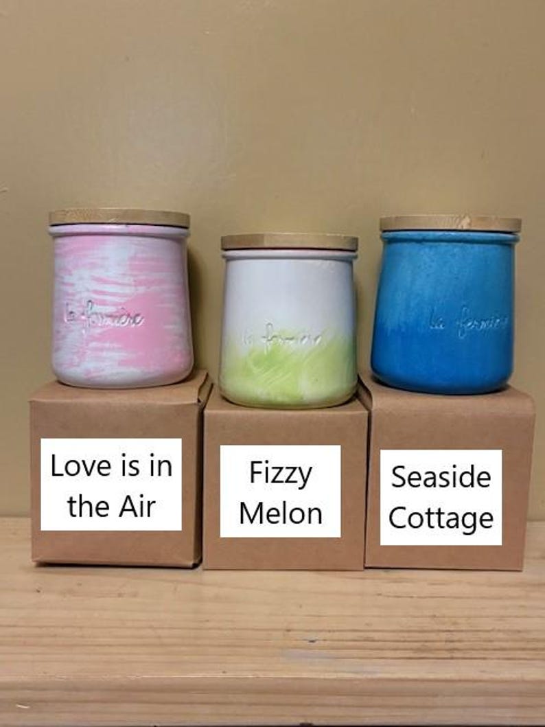 May include: Three ceramic jars with wooden lids, each featuring the embossed text la fermiere. The left jar has pink and white brushstrokes, the center jar is white with a green base, and the right jar is solid blue. Each sits on a brown box.