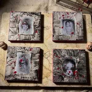 May include: Four square wall art pieces featuring black and white portraits of figures in textured, distressed frames with red paint splatters. The set includes two doll heads and several bone-like objects arranged on a marble-patterned base.