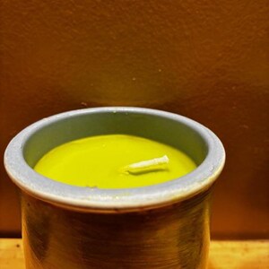 May include: A cylindrical ceramic container with a textured, dark-toned exterior and a light blue rim, filled with smooth, lime-green wax. A single white cotton wick is centered in the wax, protruding slightly from the surface of the candle.