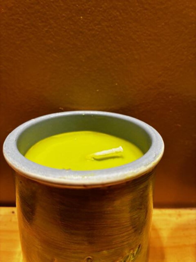 May include: A cylindrical ceramic container with a textured, dark-toned exterior and a light blue rim, filled with smooth, lime-green wax. A single white cotton wick is centered in the wax, protruding slightly from the surface of the candle.