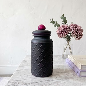 May include: A black decorative jar with a diamond pattern, a purple rim, and a magenta sphere on top. The jar sits on a marble table next to a vase of pink flowers and two books.