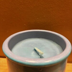 May include: A cylindrical candle in a light blue ceramic container with a smooth, matte finish. The wax inside is a matching light blue color, featuring a single centered white cotton wick. The rim of the container is a slightly darker blue tone.