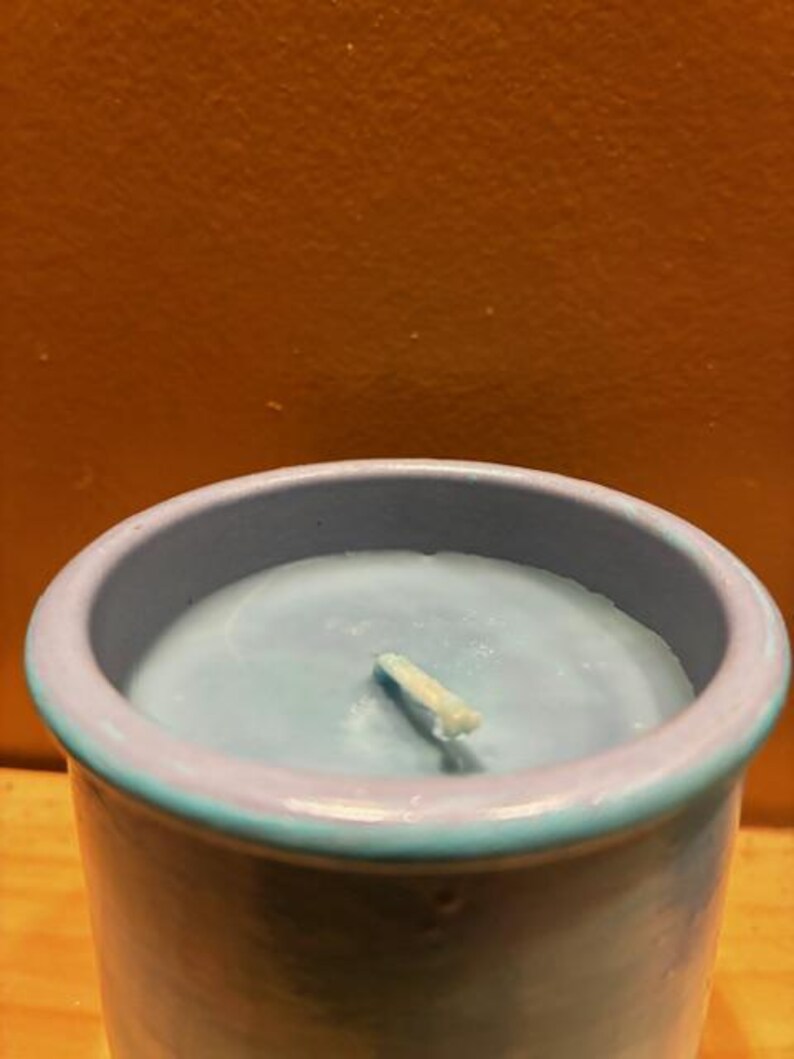 May include: A cylindrical candle in a light blue ceramic container with a smooth, matte finish. The wax inside is a matching light blue color, featuring a single centered white cotton wick. The rim of the container is a slightly darker blue tone.