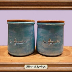 May include: Two cylindrical ceramic jars with a mottled turquoise and blue finish, each featuring a wooden lid and the embossed text "la fermi&egrave;re" on the front. The jars are displayed within a brown wooden frame with a label reading "Mineral Springs".