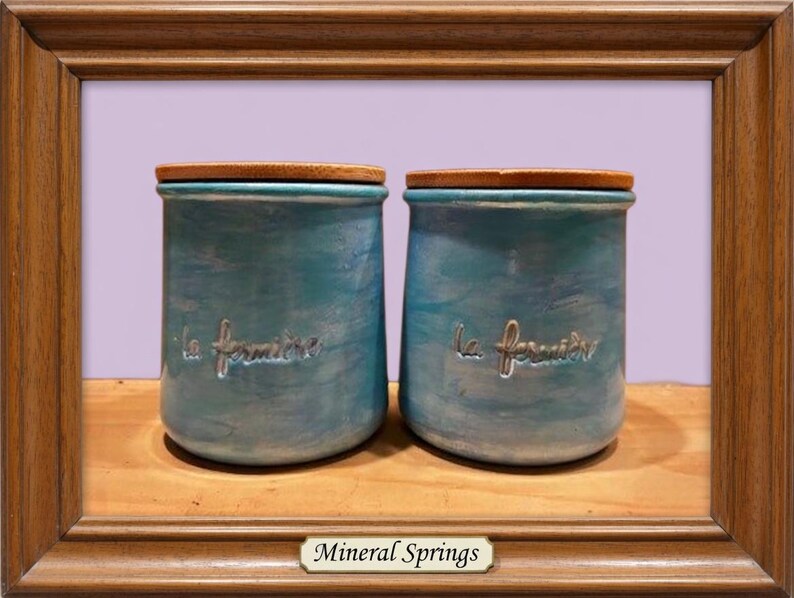 May include: Two cylindrical ceramic jars with a mottled turquoise and blue finish, each featuring a wooden lid and the embossed text "la fermi&egrave;re" on the front. The jars are displayed within a brown wooden frame with a label reading "Mineral Springs".