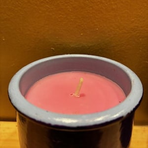 May include: A cylindrical candle container with a dark blue exterior and a light blue rim, filled with smooth pink wax. A single, centered, off-white cotton wick protrudes from the surface of the wax, which is set within the ceramic vessel.