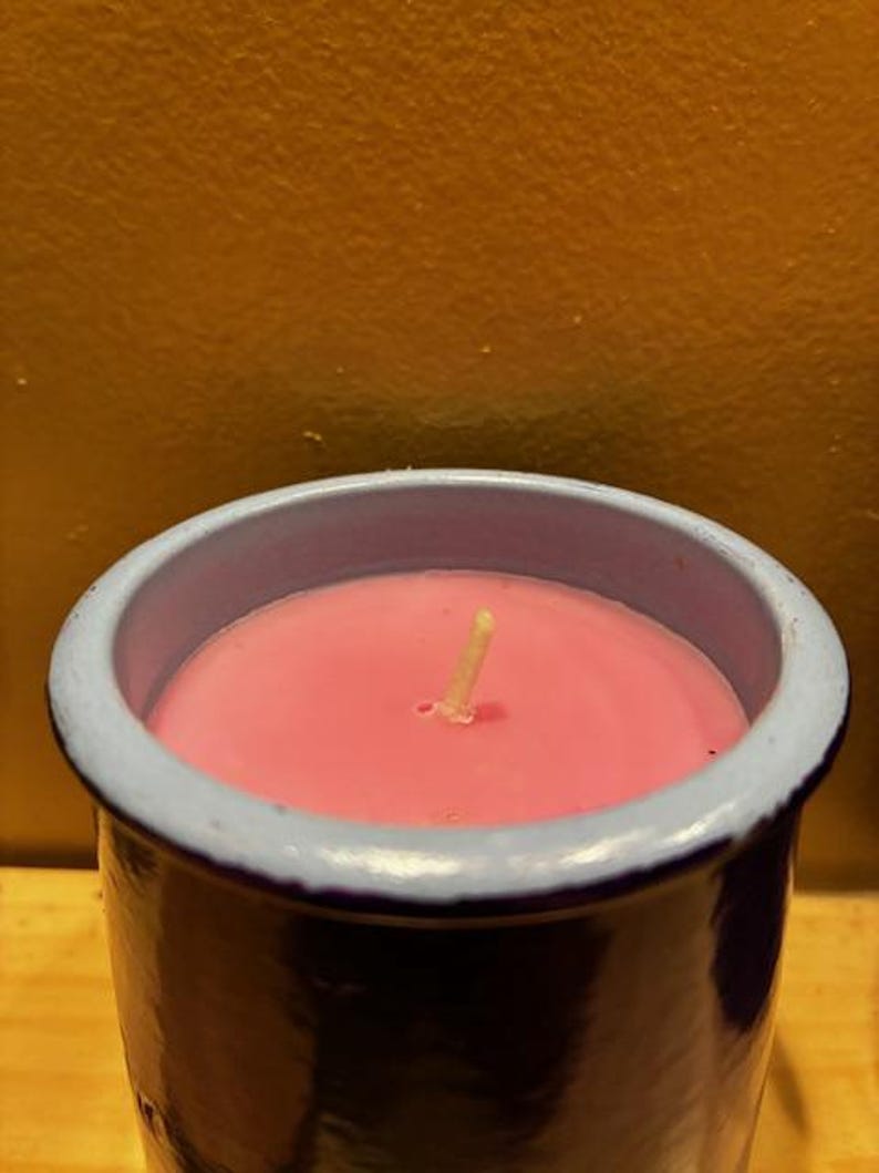 May include: A cylindrical candle container with a dark blue exterior and a light blue rim, filled with smooth pink wax. A single, centered, off-white cotton wick protrudes from the surface of the wax, which is set within the ceramic vessel.