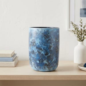 May include: A cylindrical ceramic vase featuring a mottled, multi-tonal blue and dark charcoal finish with a textured, sponge-painted appearance. The vessel has a smooth, rounded base and a wide, open top with a dark interior rim.