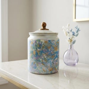 May include: A cylindrical ceramic canister featuring a marbled paint finish in shades of blue, gold, and white. The container includes a matching lid topped with a small, rounded, metallic gold-toned knob handle, creating a decorative storage vessel.