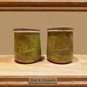 May include: Two cylindrical jars with wooden lids, featuring a textured green and brown painted finish. Each jar has the embossed text "la famille" on the front. The jars are displayed within a wooden frame with a plaque reading "Pear & Citronella".