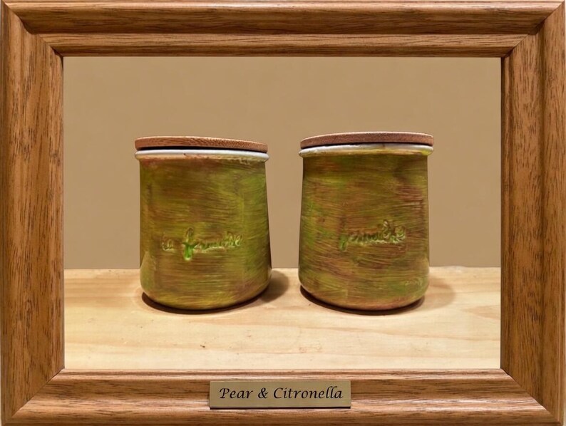 May include: Two cylindrical jars with wooden lids, featuring a textured green and brown painted finish. Each jar has the embossed text "la famille" on the front. The jars are displayed within a wooden frame with a plaque reading "Pear & Citronella".