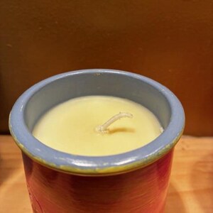 May include: A cylindrical candle in a ceramic container with a light blue rim and a red and orange painted body. The candle wax is pale yellow with a single centered white wick protruding from the surface. The container has a smooth finish.