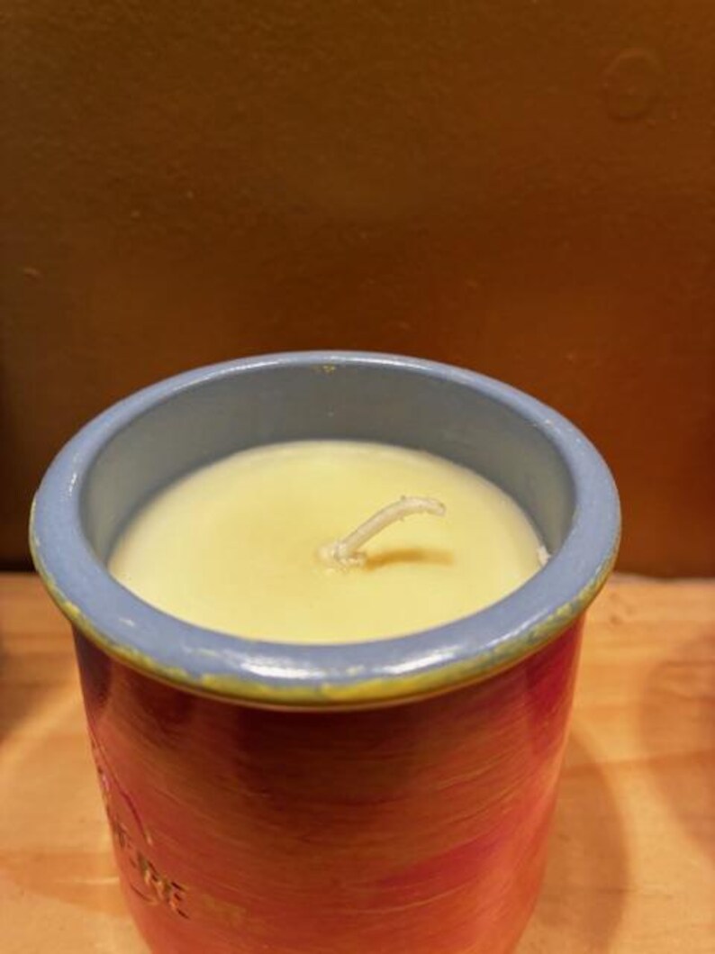 May include: A cylindrical candle in a ceramic container with a light blue rim and a red and orange painted body. The candle wax is pale yellow with a single centered white wick protruding from the surface. The container has a smooth finish.