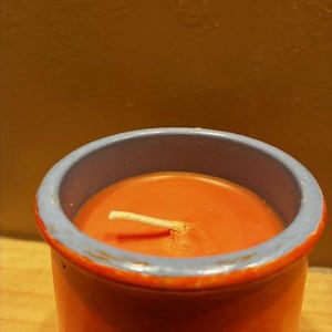 May include: A cylindrical candle in a burnt orange ceramic container with a light blue rim. The wax inside is a matching burnt orange color with a single white cotton wick centered in the middle of the vessel, extending slightly above the surface.