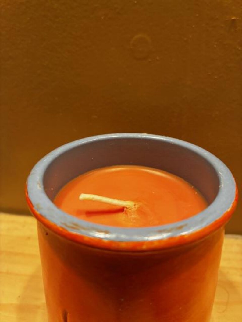May include: A cylindrical candle in a burnt orange ceramic container with a light blue rim. The wax inside is a matching burnt orange color with a single white cotton wick centered in the middle of the vessel, extending slightly above the surface.