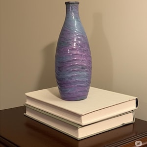 May include: A tall, decorative vase with a textured surface, featuring a blend of purple and blue hues. The vase is placed on top of two books with cream-colored covers, creating a layered display. The vase has a narrow neck and a rounded body.