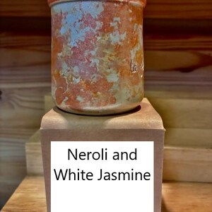 May include: A cylindrical ceramic jar with a mottled orange and white finish, topped with a flat wooden lid. The jar sits on a square tan base featuring a white label with the black printed text Neroli and White Jasmine.