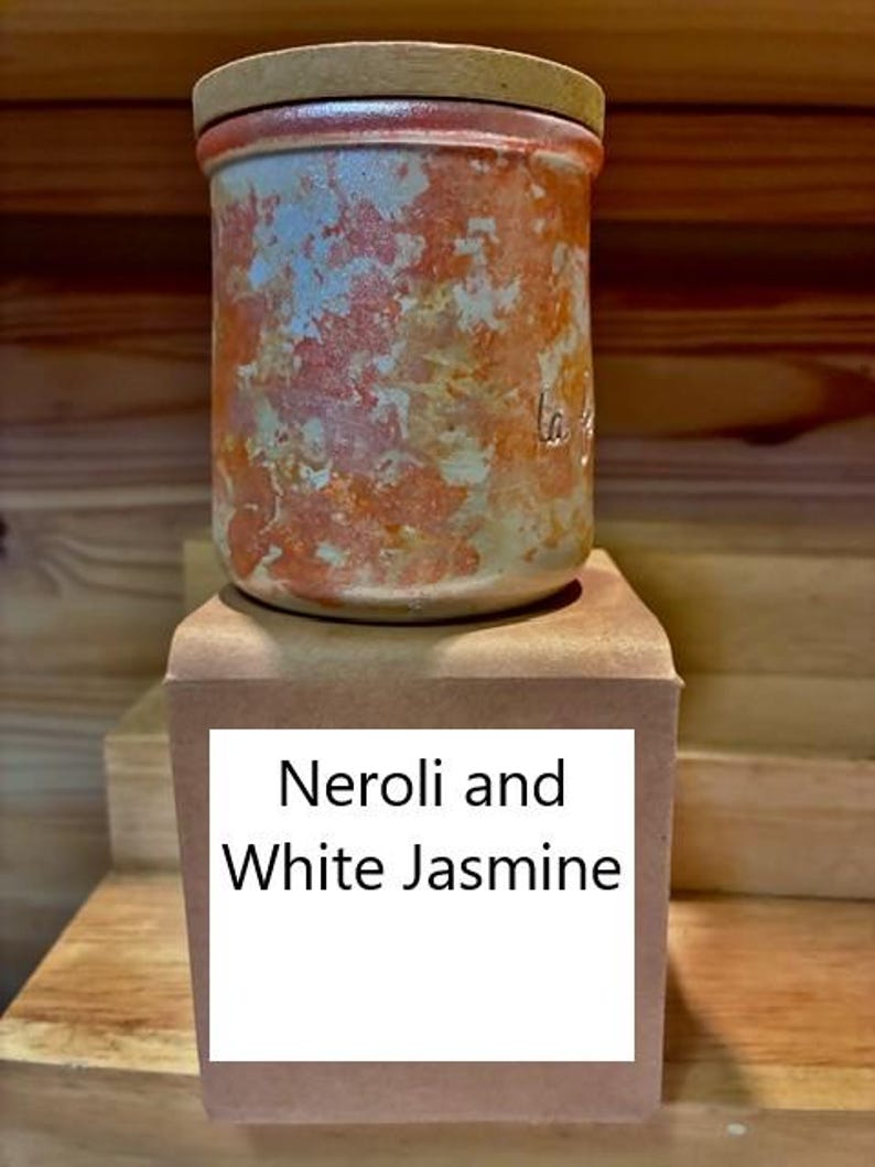 May include: A cylindrical ceramic jar with a mottled orange and white finish, topped with a flat wooden lid. The jar sits on a square tan base featuring a white label with the black printed text Neroli and White Jasmine.