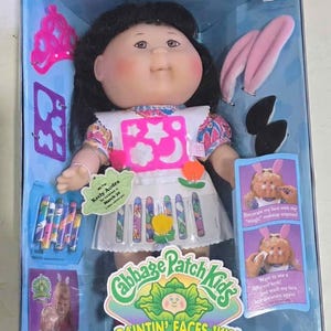 May include: A Cabbage Patch Kids Paintin' Faces Kid doll in a box. The doll has black hair, a white dress with colorful crayon designs, and a tag that reads "Keely Audra." Accessories include a pink crown and bunny ears.