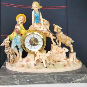 May include: A decorative clock featuring a pastoral scene with two children, a dog, goats, and sheep. The clock face is gold-toned with Roman numerals. The base is marble.