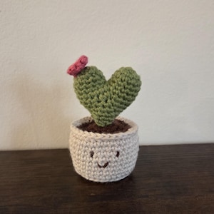 May include: A crocheted cactus in a small pot. The cactus is heart-shaped and green, with a small pink flower. The pot is light gray with a smiling face. The cactus is a charming handmade item.