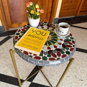 May include: A mosaic tile table with a gold frame. The table features a colourful geometric design. A book titled "YOU are a BADASS" and a cup of coffee are on the table, alongside a small potted plant with flowers.