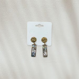 May include: A pair of gold-toned earrings with a textured, circular top and a rectangular, iridescent bottom. The earrings are displayed on a white earring card. The earrings have a post-back closure.