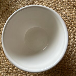May include: A white ceramic bowl with a rounded shape. The bowl is empty and sits on a woven, tan-colored mat. The bowl's interior is smooth and reflective, contrasting with the textured background.