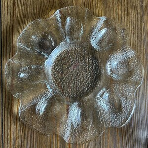 May include: A clear, textured glass serving dish with a flower-like shape. The dish has a central, slightly raised area and several petal-shaped compartments around the edge. The glass has a slightly amber hue.