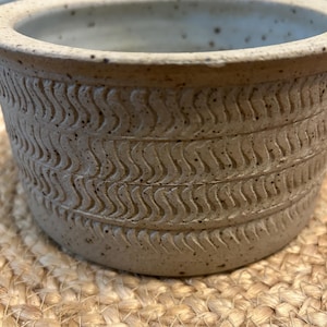 May include: A beige ceramic pot with a textured, wavy pattern around the exterior. The pot has a wide, open top and sits on a woven, tan surface. The pot's design features a series of carved, repeating wave-like lines.
