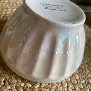 May include: Iridescent white bowl with a ribbed design. The bowl has a pearlescent finish that reflects light, creating a shimmering effect. The bottom of the bowl has text that reads "by ANTHROPOLOGIE". The bowl is sitting on a woven surface.
