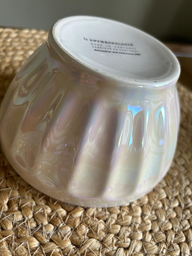 May include: Iridescent white bowl with a ribbed design. The bowl has a pearlescent finish that reflects light, creating a shimmering effect. The bottom of the bowl has text that reads "by ANTHROPOLOGIE". The bowl is sitting on a woven surface.