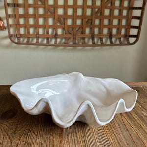May include: A white ceramic bowl shaped like a giant clam shell. The bowl features a glossy, smooth finish with deep, undulating curves and a wide, open center. The edges are scalloped and irregular, mimicking the natural form of a mollusk shell.