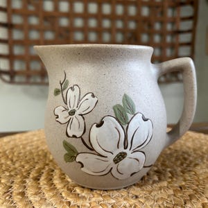 May include: A beige ceramic pitcher with a handle, decorated with white dogwood flowers and green leaves. The pitcher is sitting on a woven mat.