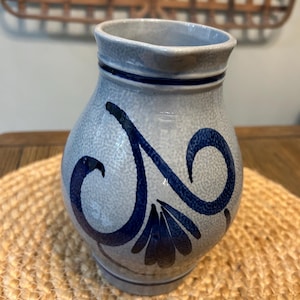 May include: A blue and gray ceramic pitcher with a bulbous body and a narrow neck. The pitcher is decorated with dark blue floral designs. The pitcher is sitting on a woven mat.