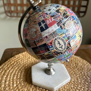 May include: A decorative globe featuring a collage of vintage travel stickers and labels. The globe is supported by a silver-toned metal stand and a white marble hexagonal base. The globe's surface is covered in colorful travel-themed imagery.