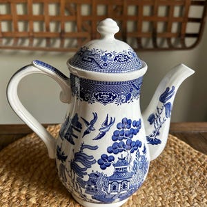 May include: White ceramic teapot with a blue Willow pattern design. The teapot features a lid, handle, and spout. The blue design includes pagodas, trees, birds, and other traditional elements. The teapot is sitting on a woven mat.