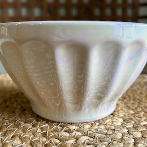 May include: A pearlescent white ceramic bowl with a scalloped design. The bowl has a glossy finish and sits on a woven surface. The bowl's shape is round with a slightly flared rim, suitable for serving food.