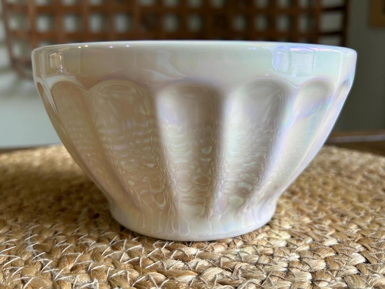May include: A pearlescent white ceramic bowl with a scalloped design. The bowl has a glossy finish and sits on a woven surface. The bowl's shape is round with a slightly flared rim, suitable for serving food.