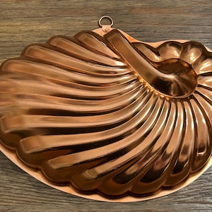 May include: A copper-colored, seashell-shaped mold with a handle and hanging loop. The mold has a detailed, ridged design and a shiny finish, perfect for baking decorative cakes or desserts.