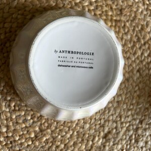 May include: A white, iridescent bowl with a textured, geometric design. The bottom of the bowl features text that reads "by ANTHROPOLOGIE", "MADE IN PORTUGAL", "FABRIQU&Eacute; AU PORTUGAL", and "dishwasher and microwave safe".