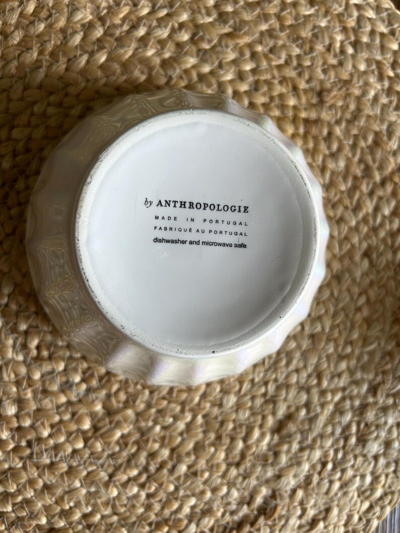 May include: A white, iridescent bowl with a textured, geometric design. The bottom of the bowl features text that reads "by ANTHROPOLOGIE", "MADE IN PORTUGAL", "FABRIQU&Eacute; AU PORTUGAL", and "dishwasher and microwave safe".