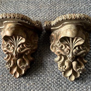 May include: Two ornate, antique-style wall shelves. Each shelf features a detailed, sculpted design with a face-like motif and floral accents. The shelves have a distressed, aged finish in shades of beige and brown.