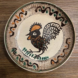 May include: A round ceramic plate with a cream-colored base featuring a central black rooster illustration with an orange crest. The rim is decorated with a repeating brown wavy line pattern, interspersed with small green and orange accents.